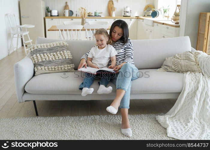 Mom and child are reading story together sitting on sofa. Caucasian mother is teaching her little daughter and hugs her. Handsome woman and girl together at home. Concept of parenthood and study.. Mom and child are reading story together sitting on sofa. Parenthood, study and support.