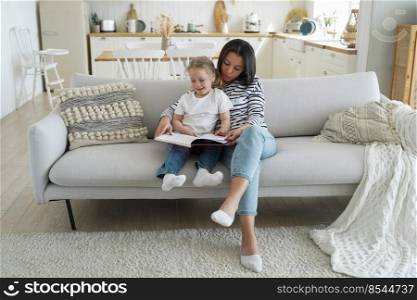 Mom and child are reading story together sitting on sofa. Caucasian mother is teaching her little daughter and hugs her. Handsome woman and girl together at home. Concept of parenthood and study.. Mom and child are reading story together sitting on sofa. Parenthood, study and support.