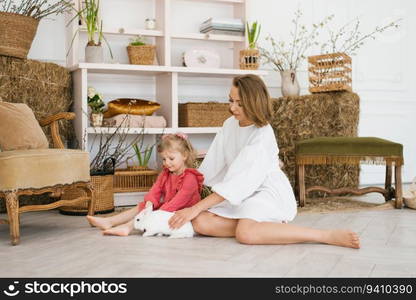 Mom and daughter play with a white rabbit, sitting on the floor in the living room, decorated with Easter