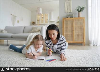 Mom and her kid are drawing together. Young caucasian woman and little daughter lying on floor together and coloring with markers. Happy family has good time at home. Childhood and parenthood concept.. Young caucasian woman and little daughter lying on floor together and coloring with markers