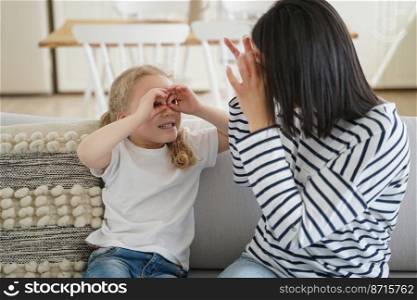 Mom and kid little daughter makes funny faces, having fun, playing together, sitting on sofa at home. Mother and small girl showing glasses by hands, looking to each other, fooling around.. Mom and kid little daughter make funny faces, having fun, playing together, sitting on sofa at home