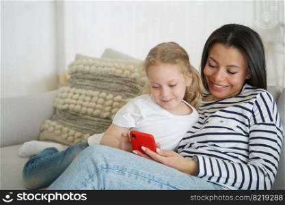 Mom and little daughter watching cartoons on smartphone, playing online game, sitting on couch. Mother and small girl use learning mobile app enjoying leisure time on sofa together.. Mom and little daughter watching cartoons on smartphone, enjoying leisure time on sofa together