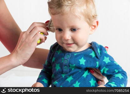 Mom combing the baby with blonde hair, while the toddler is sitting.