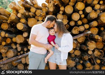 Mom, dad and their baby son are kissing in a pile of tree trunks.