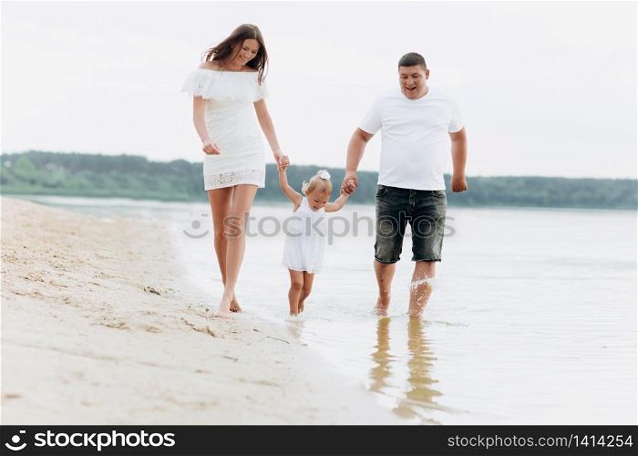 Mom, dad hugging daughter walking on the beach near lake. The concept of summer holiday. Mother&rsquo;s, father&rsquo;s, baby&rsquo;s day. Family spending time together on nature. Family look. Sun light. Mom, dad hugging daughter walking on the beach near lake. The concept of summer holiday. Mother&rsquo;s, father&rsquo;s, baby&rsquo;s day. Family spending time together on nature. Family look. Sun light.