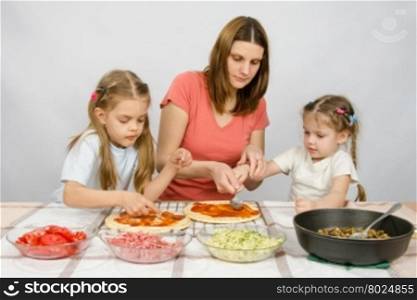 Mom helps younger daughter spread ketchup on a pizza, the eldest daughter, she is preparing a second pizza