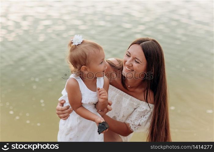 Mom hugging daughter on the beach near lake. The concept of summer holiday. Mother&rsquo;s, baby&rsquo;s day. Spending time together. Family look. Sun light. Mom hugging daughter on the beach near lake. The concept of summer holiday. Mother&rsquo;s, baby&rsquo;s day. Spending time together. Family look. Sun light.
