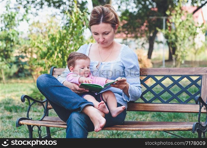 Mom reading a book and showing the pictures her little daughter sitting on bench in the garden