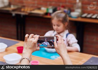 Mom recording her little daughter while taking part in baking workshop. Baking classes for children, aspiring little chefs. Learning to cook. Combining and stirring prepared ingredients. Real people, authentic situations