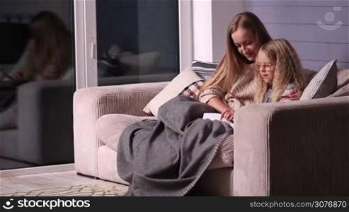 Mom teaching daughter to read at home. Cute little girl with her beautiful mother wrapped in a blanket sitting on the couch and reading a book together. Mother correcting a mistakes while her lovely daughter is learning to read.