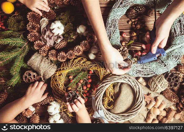 Mom with children making natural Christmas wreathes for decorate home. Top view table with hands. Nature wreath making