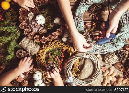 Mom with children making natural Christmas wreathes for decorate home. Top view table with hands. Nature wreath making
