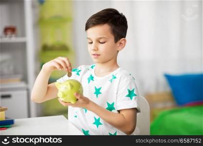 money, finances, childhood and people concept - little boy putting coin into piggy bank at home. little boy putting coin into piggy bank at home