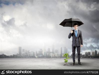 Money making. Young businessman with umbrella protecting sprout growing in desert