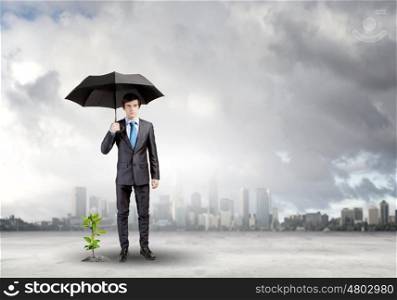 Money making. Young businessman with umbrella protecting sprout growing in desert