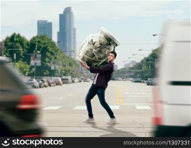 Money saving concept, young man trying to carry a big glass jar full of dollars. Cash economy addiction, home bank. Man trying to carry a glass jar full of dollars