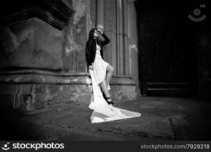 Monochrome full length portrait of woman in long white dress leaning against stone wall