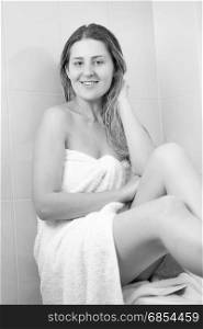 Monochrome image of beautiful woman with long hair covered in white towel sitting on bath