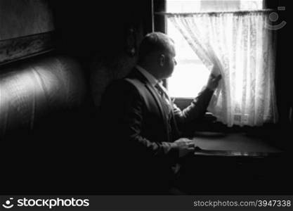 Monochrome portrait of elegant man sitting in train coupe and looking out of window