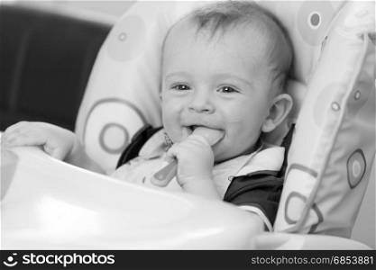 Monochrome portrait of smiling playful baby eating from spoon in highchair