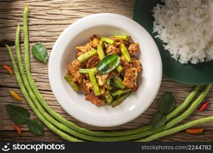 Moo Pad Prik Gaeng, Thai food, stir fried pork with red curry paste, bergamot leaves and yardlong beans beside streamed rice on rustic natural wood texture background, top view