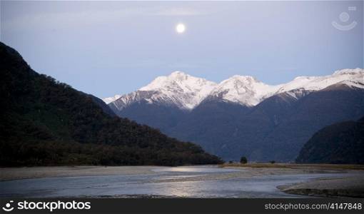 Moon rises over snow capped mountains with river in foreground