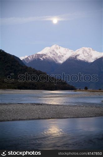 Moon rises over snow capped mountains with river in foreground
