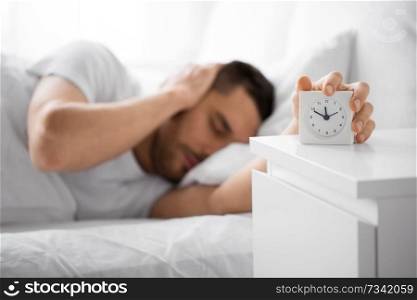 morning and people concept - close up of sleepy young man in bed reaching for alarm clock on bedside table at home. close up of man in bed reaching for alarm clock