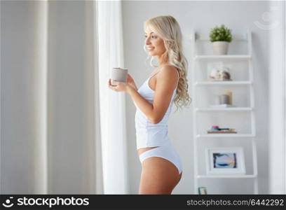 morning and people concept - happy young woman in white underwear with mug drinking coffee at home window. woman in underwear drinking coffee at home window