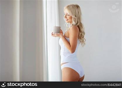 morning and people concept - happy young woman in white underwear with mug drinking coffee at home window. woman in underwear drinking coffee at home window