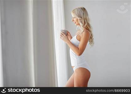 morning and people concept - happy young woman in white underwear with mug drinking coffee at home window. woman in underwear drinking coffee at home window. woman in underwear drinking coffee at home window