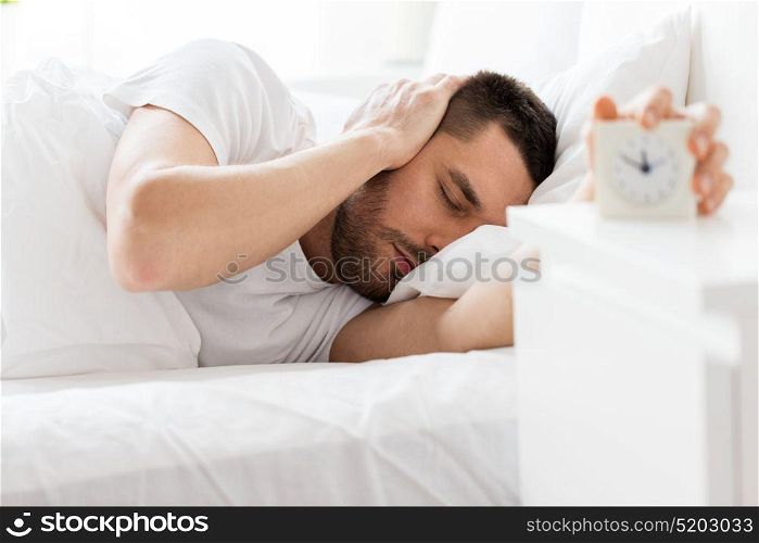 morning and people concept - sleepy young man in bed reaching for alarm clock at home. young man in bed reaching for alarm clock