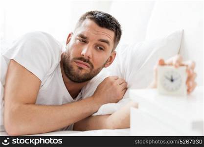morning and people concept - sleepy young man in bed reaching for alarm clock at home. young man in bed reaching for alarm clock