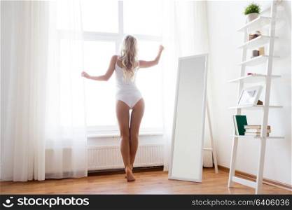 morning and people concept - young woman in white underwear looking through window at home. woman in underwear at window in morning