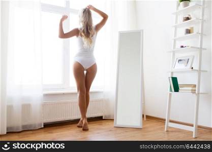 morning and people concept - young woman in white underwear looking through window at home. woman in underwear at window in morning