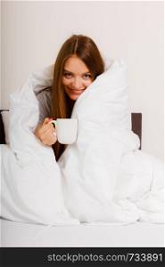 Morning, awakening drink concept. Beautiful smiling young woman holding a cup of hot coffee or tea in bed indoor.. Smiling woman holding cup of drink in bed
