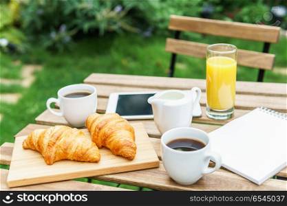 Morning Breakfast In Green Garden With French Croissant, Coffee Cup, Orange Juice, Tablet and Notes Book On Wooden Table