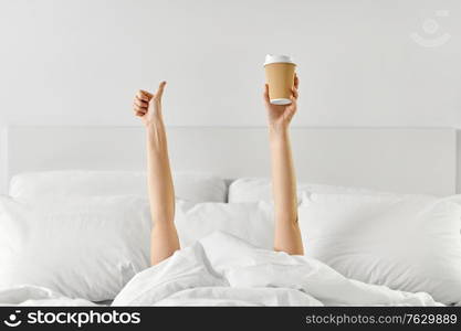 morning, comfort and people concept - young woman with cup of takeaway coffee lying in bed and showing thumbs up at home bedroom. woman with coffee lying in bed showing thumbs up