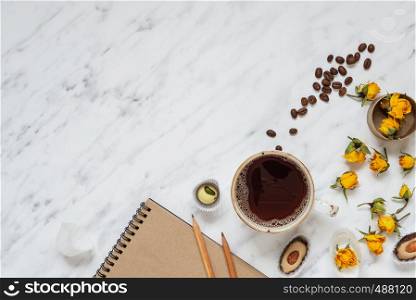 Morning composition with a cup of black coffee, chocolate candies, dry yellow rose flowers and a notebook on a marble surface with space for text, top view