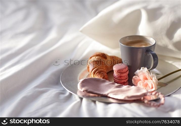 morning, hygge and breakfast concept - croissant, cup of coffee, macaroons and eye sleeping mask on plate in bed at home. croissant, coffee and eye sleeping mask in bed