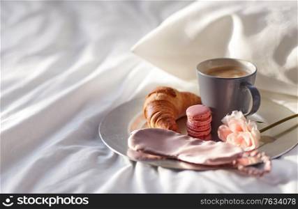 morning, hygge and breakfast concept - croissant, cup of coffee, macaroons and eye sleeping mask on plate in bed at home. croissant, coffee and eye sleeping mask in bed