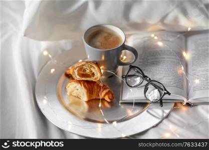 morning, hygge and breakfast concept - croissants, cup of coffee, book and glasses in bed at home. croissants, cup of coffee, book and glasses in bed