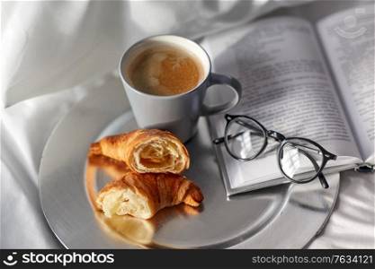 morning, hygge and breakfast concept - croissants, cup of coffee, book and glasses in bed at home. croissants, cup of coffee and book in bed at home
