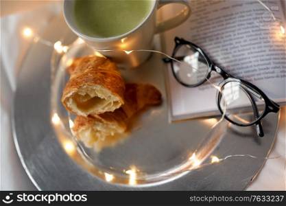 morning, hygge and breakfast concept - croissants, cup of matcha tea book and glasses in bed at home. croissants, matcha tea, book and glasses in bed