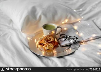 morning, hygge and breakfast concept - croissants, cup of matcha tea book and glasses in bed at home. croissants, matcha tea, book and glasses in bed