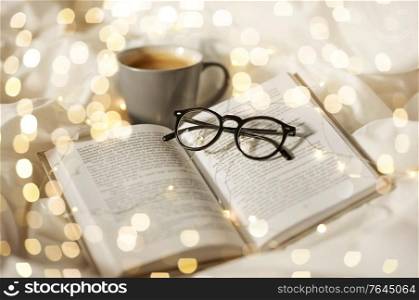 morning, hygge and breakfast concept - cup of coffee, book, glasses and garland lights in bed at home. cup of coffee, book, glasses and garland in bed