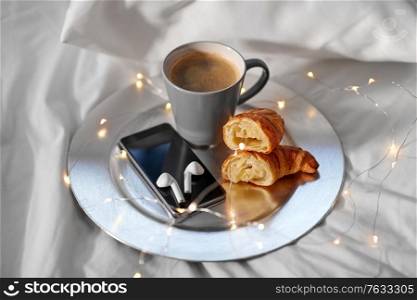 morning, hygge and breakfast concept - smartphone, wireless earphones, cup of coffee and croissants with garland lights on plate in bed at home. smartphone, earphones, coffee and croissant in bed