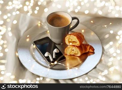 morning, hygge and breakfast concept - smartphone, wireless earphones, cup of coffee and croissants with garland lights on plate in bed at home. smartphone, earphones, coffee and croissant in bed