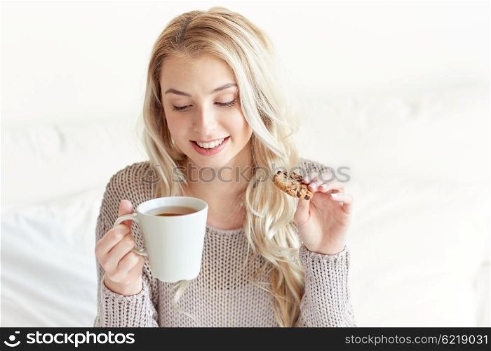 morning, leisure and people concept - happy young woman with cup of tea eating cookie in bed at home bedroom