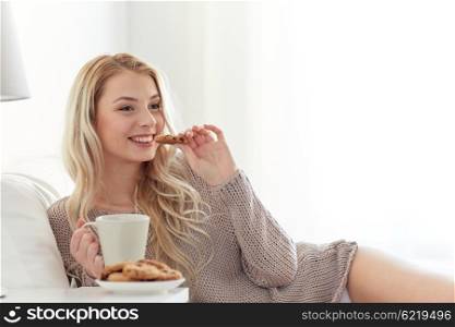 morning, leisure and people concept - happy young woman with cup of coffee or tea eating cookie in bed at home bedroom